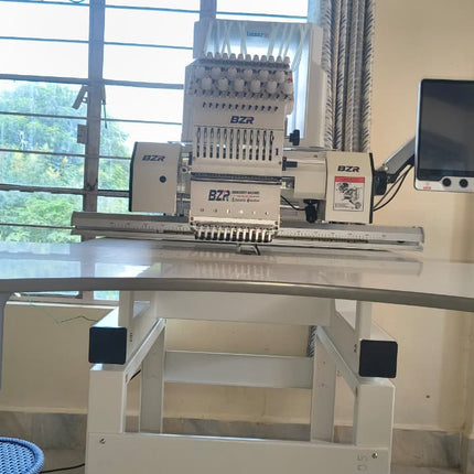 An industrial multi-head embroidery machine with a digital screen, mounted on a white adjustable work table in a room with a window.