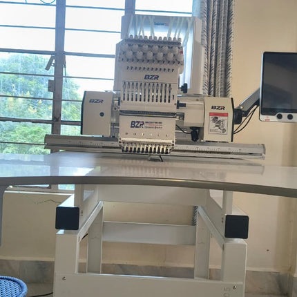 An industrial embroidery machine with a digital screen attachment, positioned on a white adjustable worktable in a room with a window.